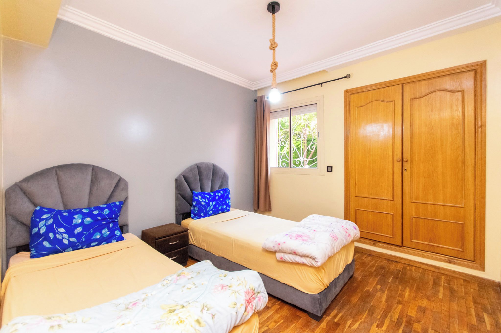 A bedroom with two beds, a wardrobe, and a window with a view of greenery.