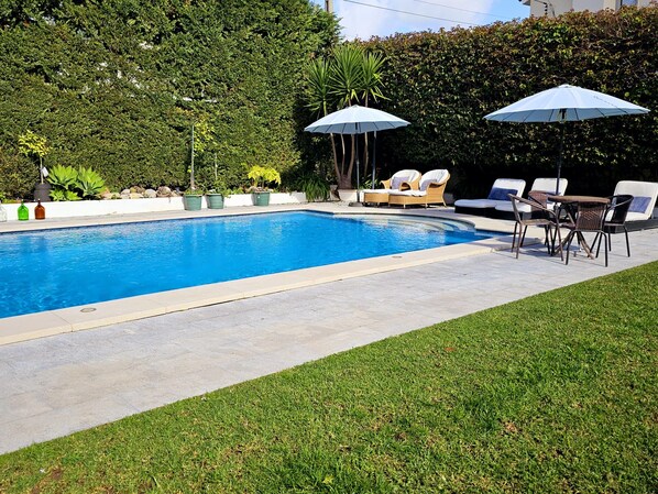 Une piscine entourée de chaises longues et de parasols, elle-même bordée d’une pelouse bien entretenue et de haies.