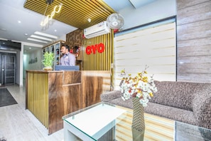 A reception area with a wooden reception desk, a sofa, a glass table, and a wall-mounted fan. - Hotel O TAJ STARS (Zirakpur)