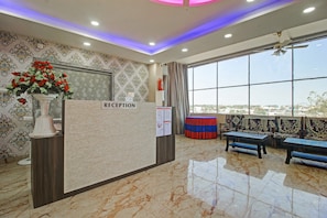 Zona de recepción con piso de mármol, un mostrador de recepción con la leyenda 'RECEPTION', un ventanal con vistas y un área de descanso con cojines estampados.