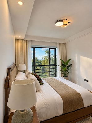 Un dormitorio con una cama grande, una mesita de noche, una lámpara, una ventana con cortinas y vistas al exterior con vegetación.