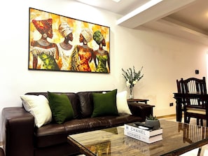 Un salon moderne avec un canapé en cuir marron, des coussins verts, une table basse en verre sur laquelle sont posés des livres et une télécommande, et un grand tableau abstrait accroché au mur.