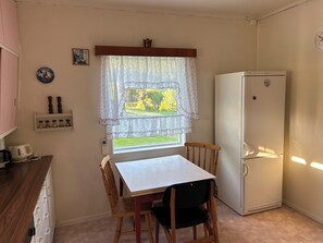 Una cuina petita amb taula i caires, un frigorífic, una finestra amb cortines i una estant mural amb objectes decoratius.