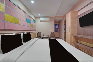 Quarto de hotel com cama, duas cadeiras, mesa com chaleira e garrafa de água, ar condicionado e TV fixada na parede.