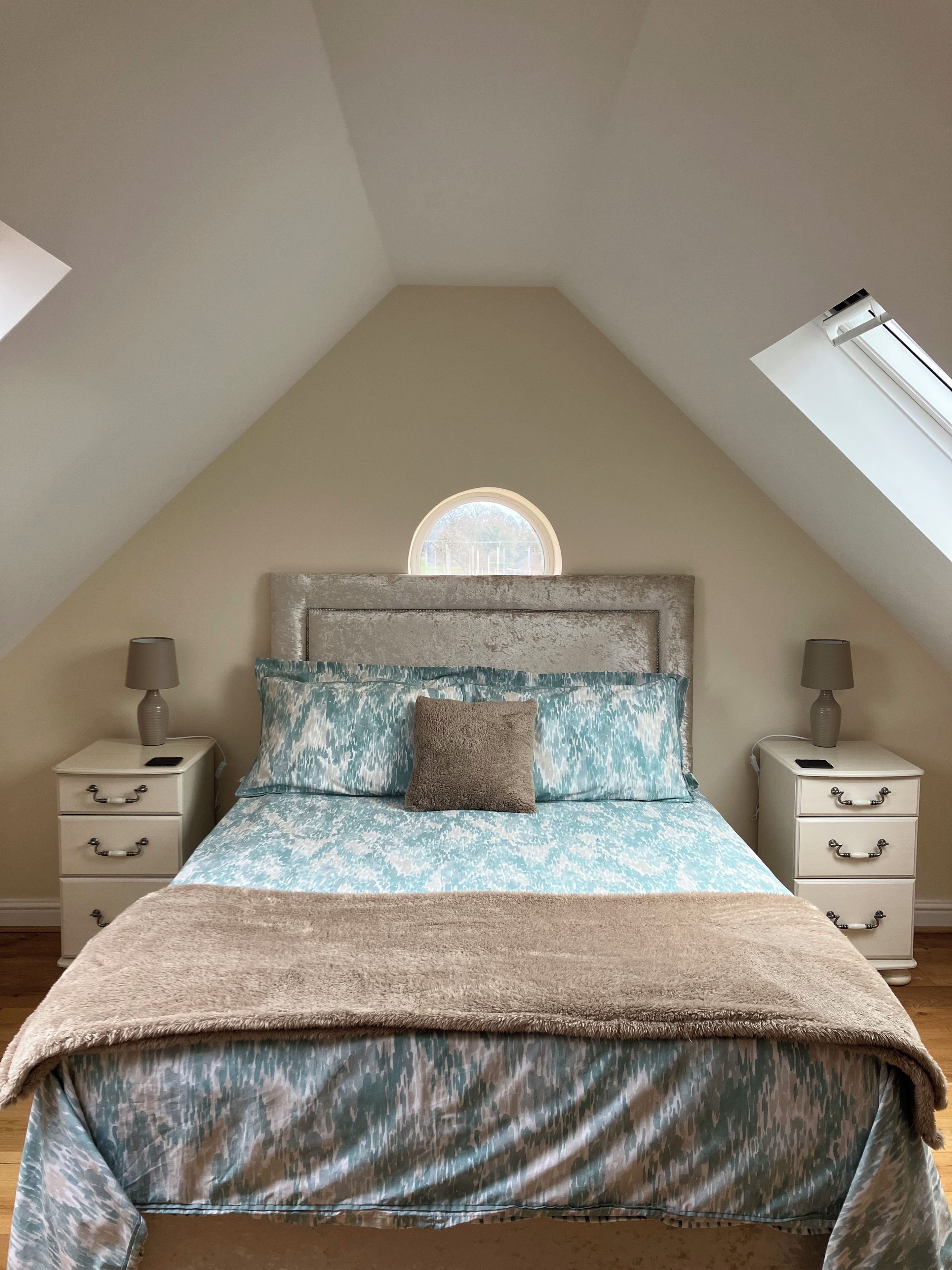 A bedroom with a sloped ceiling, a skylight, a bed with patterned bedding, two bedside tables with lamps, and a view through a window.