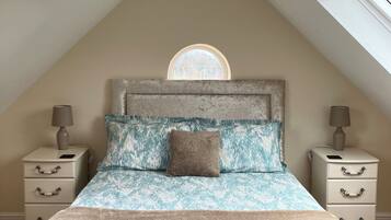 A bedroom with a sloped ceiling, a skylight, a bed with patterned bedding, two bedside tables with lamps, and a view through a window.