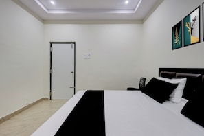Hotellihuone, jossa on sänky, yöpöytä, ovi ja seinälle kiinnitetty taideteos.