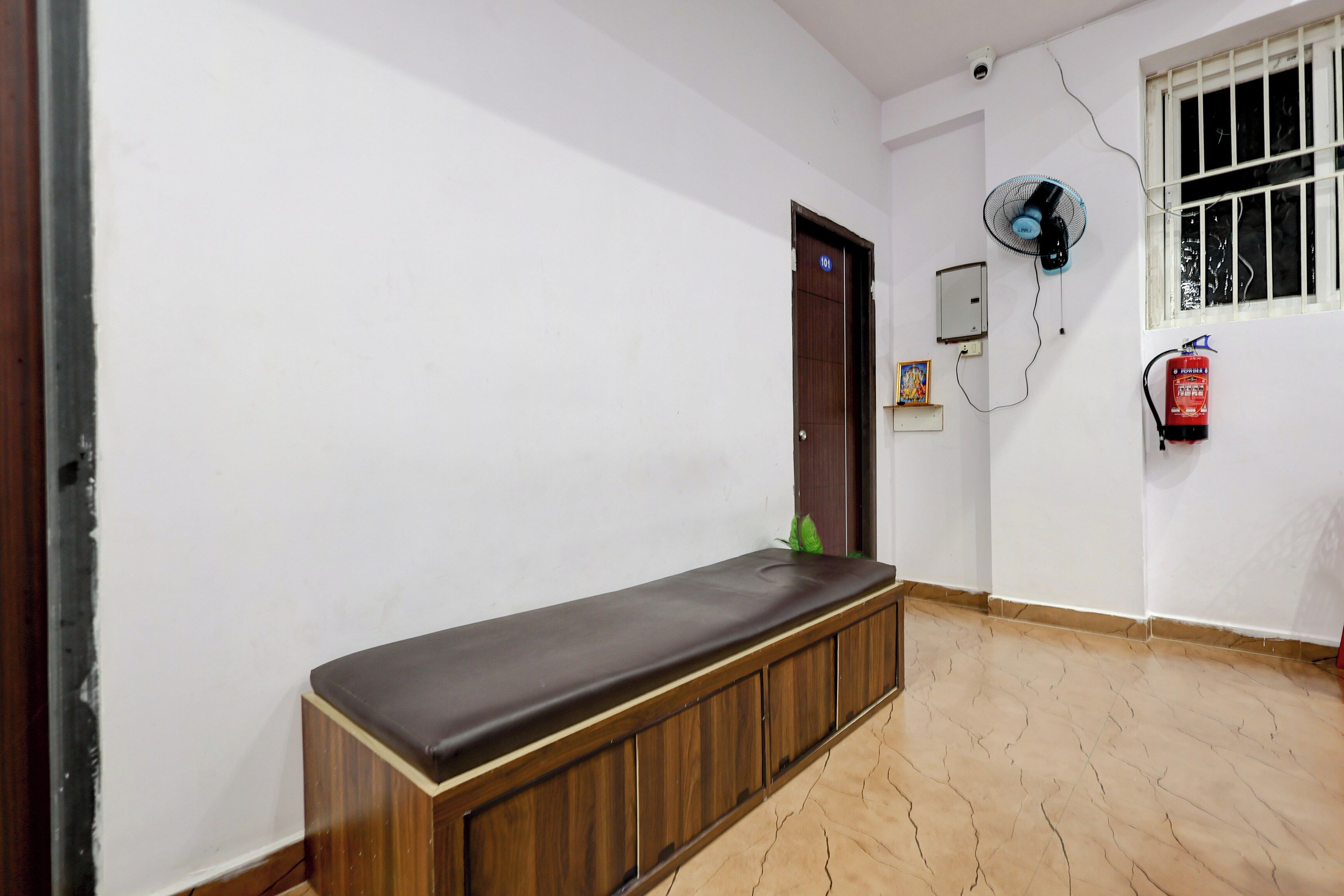A hallway with a bench, a door, a wall-mounted fan, a fire extinguisher, and a window with bars.