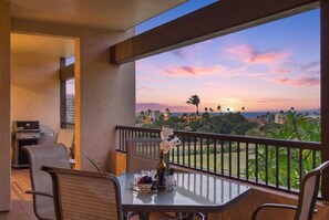 Un balcon avec une table dressée pour le dîner, un barbecue et une vue sur un coucher de soleil se reflétant sur un paysage parsemé de bâtiments et de palmiers.