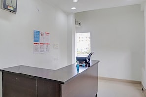 Un mostrador de recepción en el vestíbulo de un hotel, con vistas al exterior por una ventana, un interfono en la pared y algunos carteles informativos.