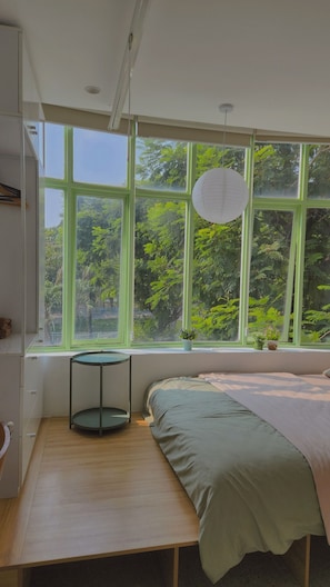Une chambre avec une grande fenêtre, un lit, une petite table et un luminaire suspendu.