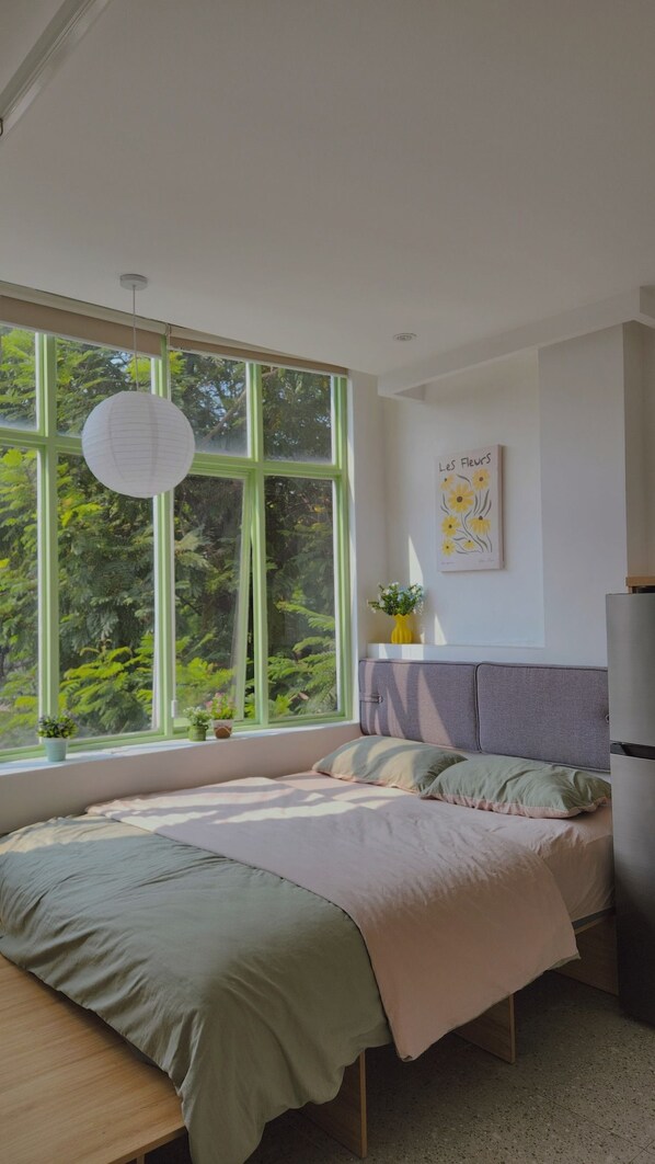 Une chambre à coucher avec un lit, une table de chevet ornée d’un vase, une fenêtre donnant sur un paysage verdoyant et un mur décoré d’un tableau représentant des fleurs.