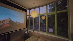 A room with a large projection screen, a bed, a small table with a plant, and a window with green frames. - D1 Heart · Parkside & Rooftop · Leafy @ Cozinema (Ben Thanh, District 1)
