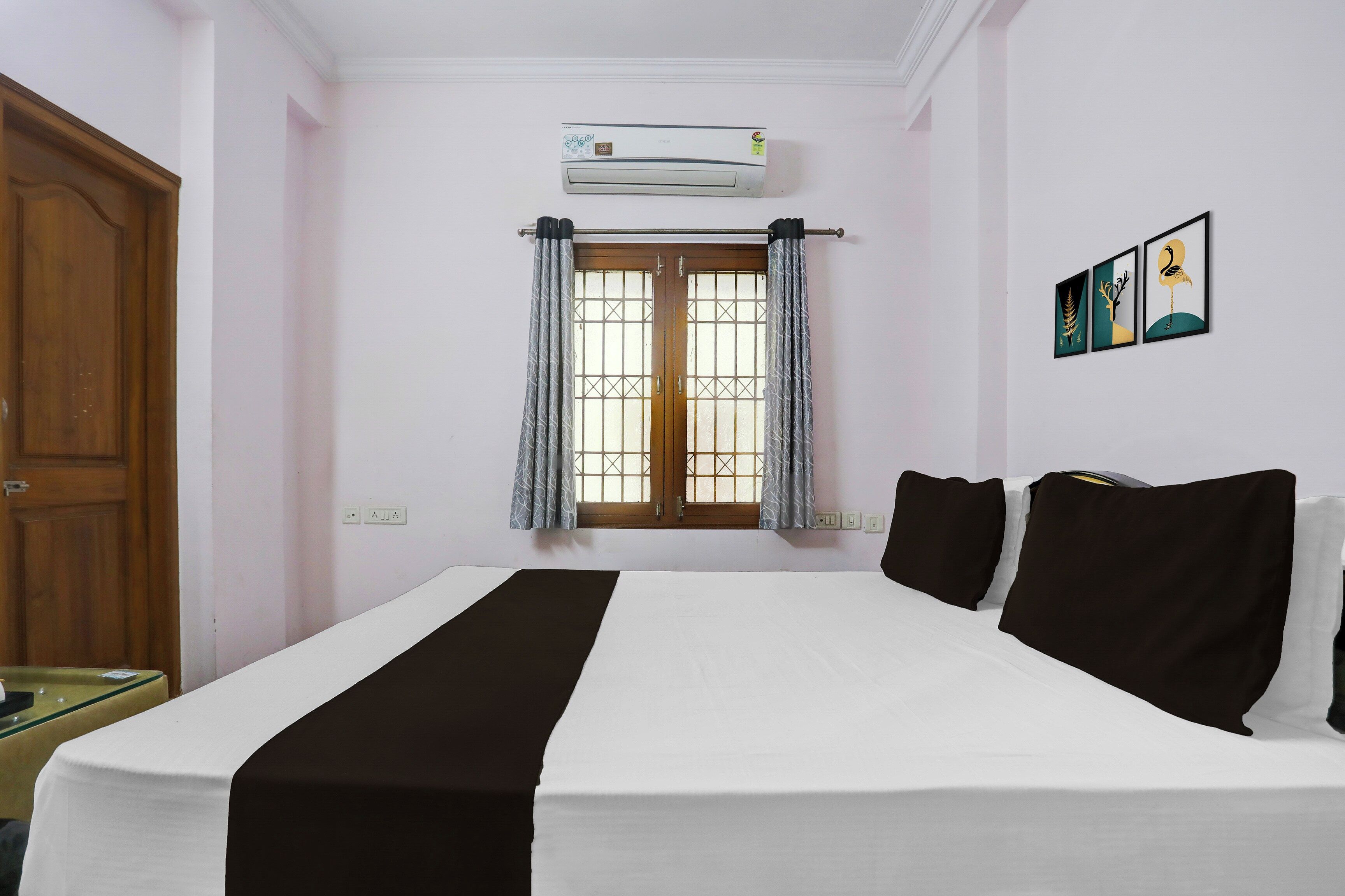 A hotel room with a bed, a window with curtains, an air conditioning unit, and a wall-mounted artwork.