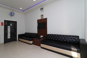Un área de recepción con un sofá negro, un mueble de madera, un televisor montado en la pared y una puerta con cámara de seguridad.