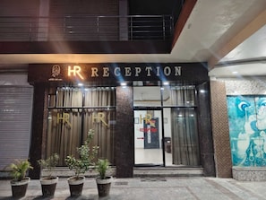 Una recepción de hotel con fachada oscura, amplios ventanales de vidrio y plantas en macetas al frente.