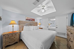 Une chambre à coucher avec un lit, deux tables de chevet avec des lampes, un ventilateur de plafond, une tête de lit en bois, une porte et un tableau accroché au mur.