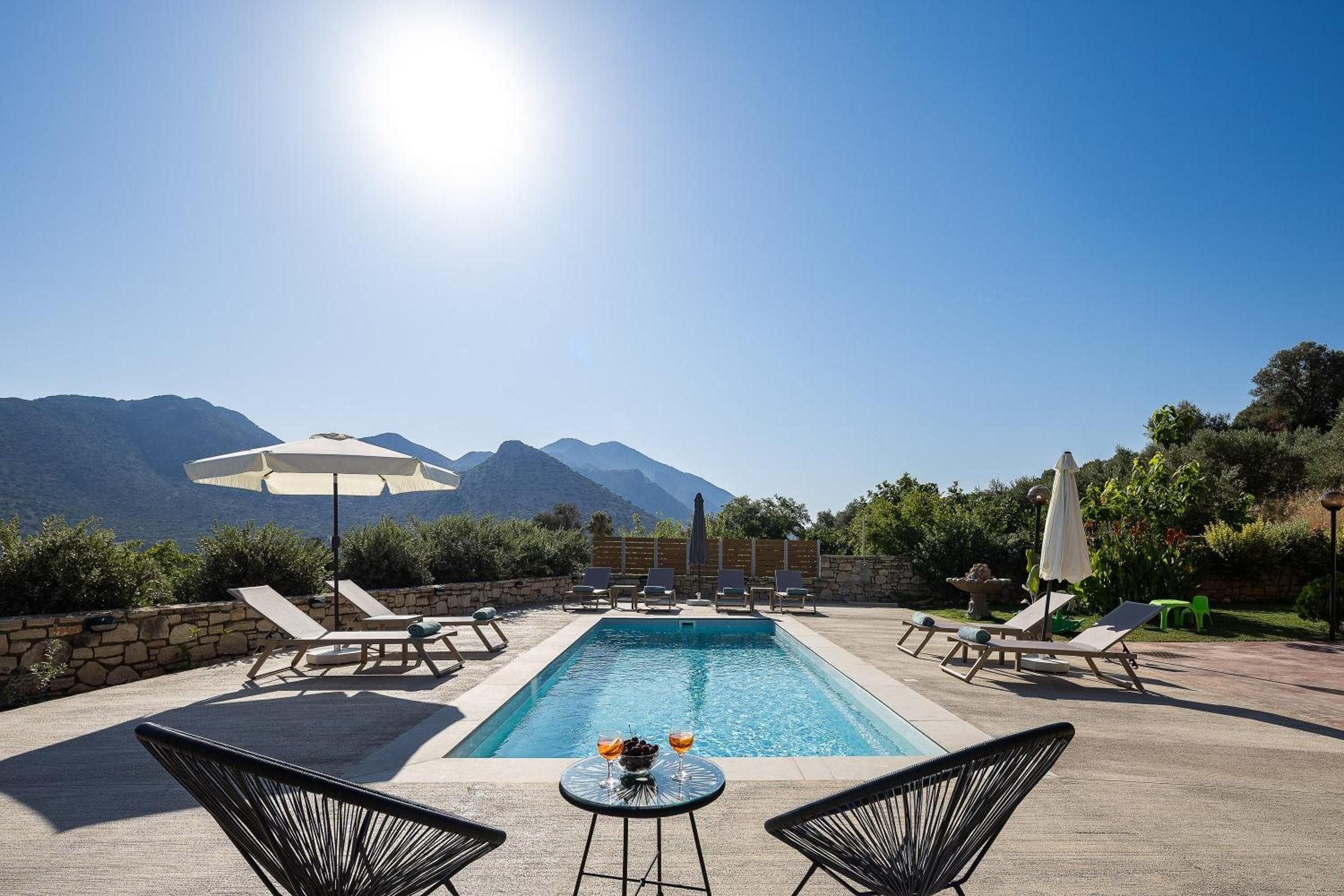 A pool with lounge chairs and umbrellas, surrounded by a stone wall and greenery, with mountains in the background.
