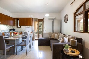 A modern living room with a sofa, a dining table with chairs, a kitchen area with cabinets, and a window with curtains. - Villa 'Leondis' with Mountain View, Wi-Fi and Air Conditioning (Μελιδόνι)