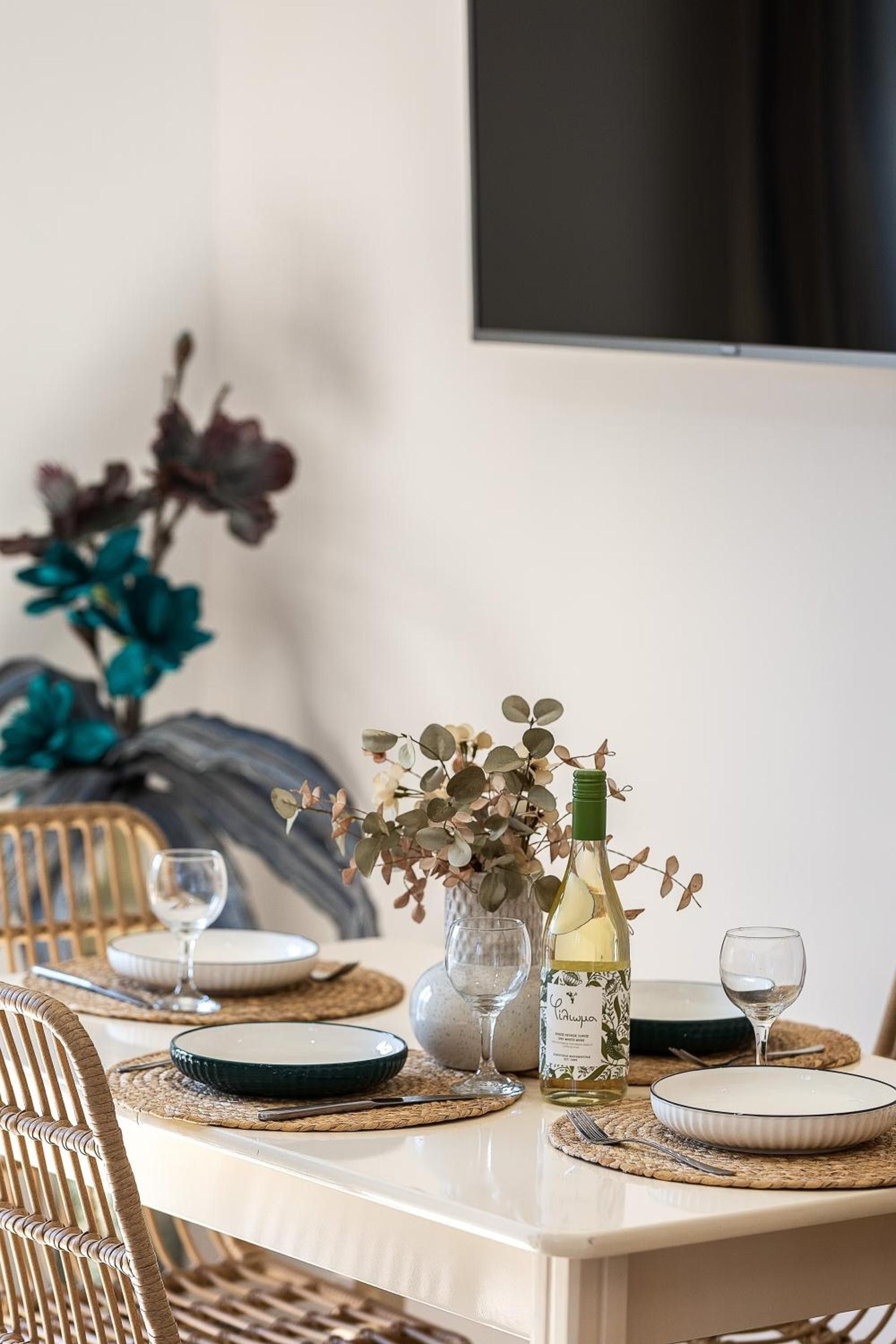 A dining table set with plates, a wine bottle, and glasses, with a decorative vase and foliage in the background.