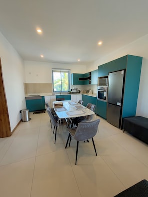 A modern kitchen with a dining area, featuring a large table, chairs, a refrigerator, and a window with blinds. - Lovely 2-bedroom apartment with lift, swimming pool in Grand Baie (Grand Baie)