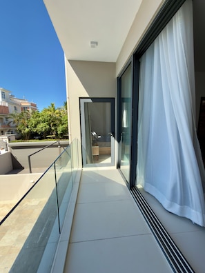 Un balcón con puerta de vidrio que da a una habitación, una cortina blanca y vistas de un edificio y árboles.