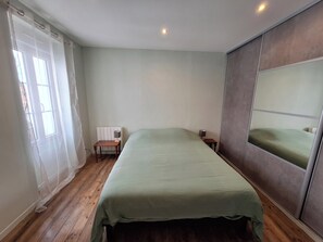 Un dormitorio con cama, mesita de noche, silla, ventana con cortinas y espejo.