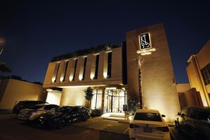 Un hotel con una facciata moderna, un cartello verticale ben visibile con il nome dell'hotel e un'area di parcheggio con diverse auto.