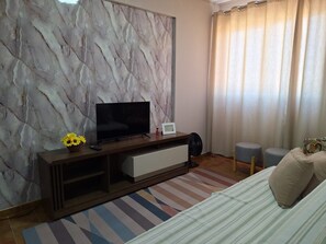 A hotel room with a marble-patterned wall, a flat-screen TV, a bedside table with a remote, a small fan, a striped rug, and a bed with pillows. - New Listing (Rondonópolis)