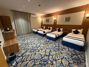 Habitación de hotel con cuatro camas dispuestas en fila, cabecera de madera, alfombra estampada y una pequeña mesita de noche de madera.