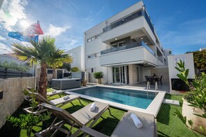 Um edifício residencial moderno de dois andares, com piscina, mobiliário de exterior e um jardim bem cuidado.