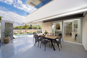 Um pátio moderno ao ar livre com piscina, mesa de jantar e cadeiras, e porta de vidro que dá para uma sala de estar.