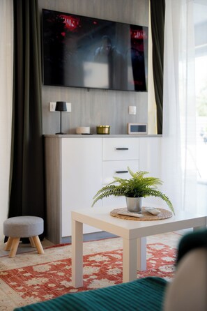 Een moderne woonkamer met een flatscreen televisie aan de wand, een klein tafeltje met een plant, een lamp en een afstandsbediening op een geweven onderzetter.