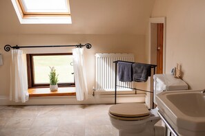 Un baño con tragaluz, una ventana con cortinas, un radiador, un inodoro y un perchero para toallas.