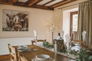 Una mesa de comedor dispuesta con platos, vasos y una botella, con un cuadro de yakas en la pared y una ventana con cortinas.