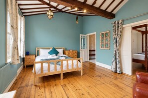 Une chambre avec un lit en bois, une table de chevet, une commode, un plancher en bois, un plafond avec des poutres apparentes et une porte donnant sur une autre pièce.