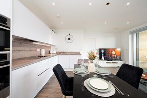 Una cocina moderna con armarios blancos, un salpicadero de madera y un rincón comedor con una mesa negra para dos.