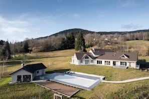 Een modern huis met twee verdiepingen en een groot terras, omgeven door een glooiend landschap en een bebost gebied.