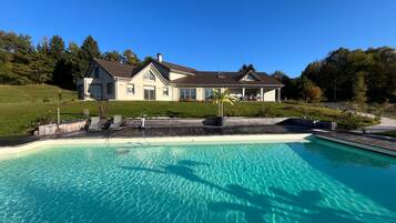 En stor swimmingpool med klart vand, en træterrasse og et hus med skråt tag i baggrunden.