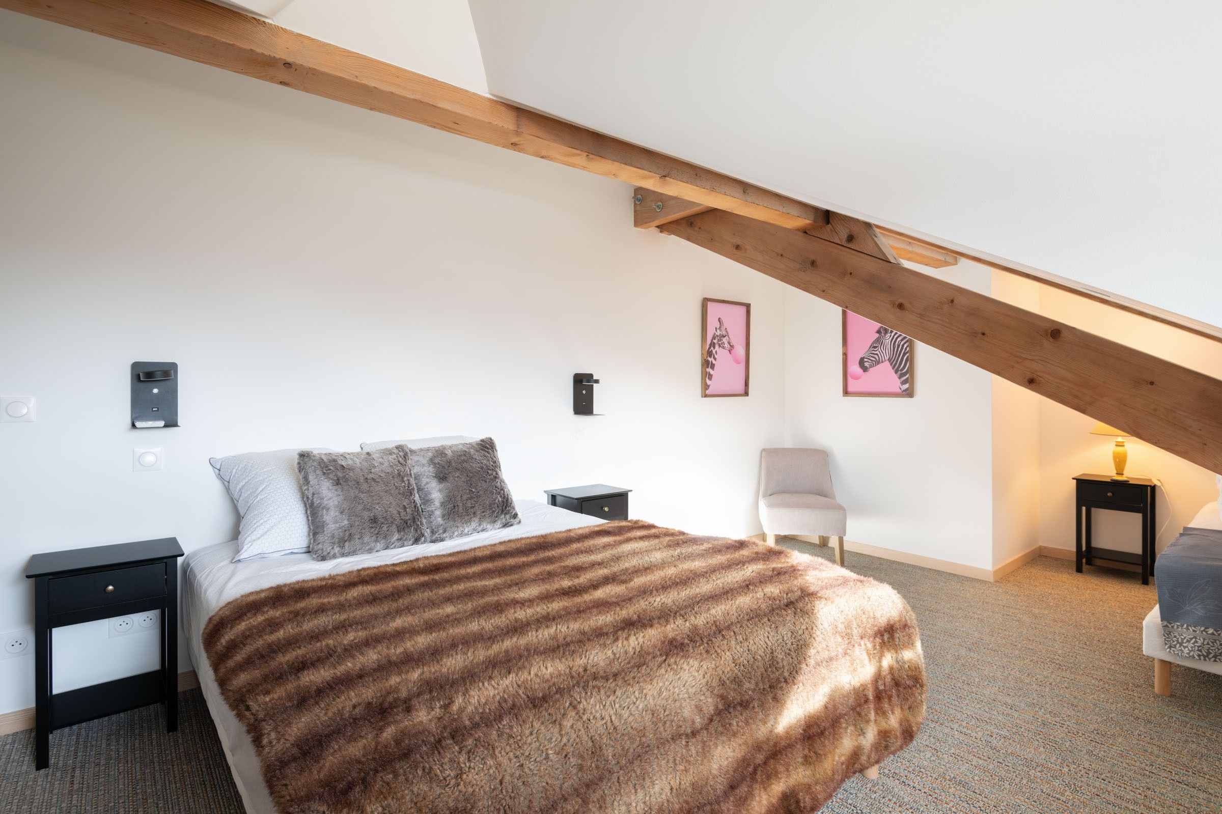 Une chambre avec un plafond en pente, un lit recouvert d’une couverture moelleuse, deux tables de chevet, une chaise et deux tableaux encadrés accrochés au mur.