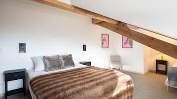 Une chambre avec un plafond en pente, un lit recouvert d’une couverture moelleuse, deux tables de chevet, une chaise et deux tableaux encadrés accrochés au mur.