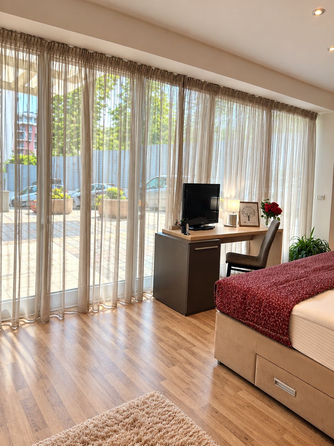 Habitación de hotel con piso de madera, un escritorio con una computadora, una silla, cama con una manta roja y un ventanal con cortinas traslúcidas.