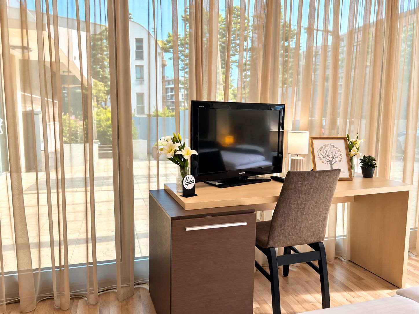 Habitación de hotel moderna con un ventanal grande, un televisor de pantalla plana sobre un escritorio de madera, una silla y una lámpara de escritorio pequeña.