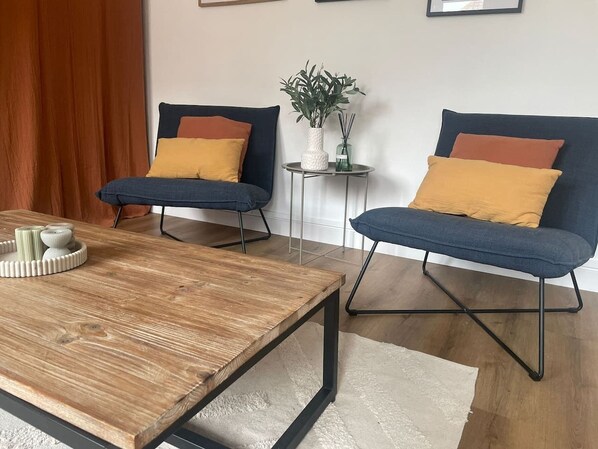 Ein Zimmer mit einem Holztisch, zwei blauen Sesseln mit senffarbenen Kissen, einem kleinen runden Tisch mit einer Pflanze und einem weißen Teppich.