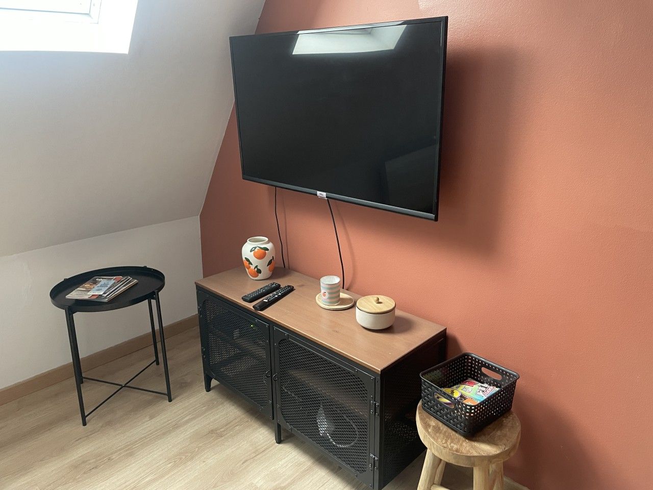 Ett rum med en platt-TV som är monterad på väggen, ett litet träskåp med en fjärrkontroll, en vas och en korg som står på en pall.