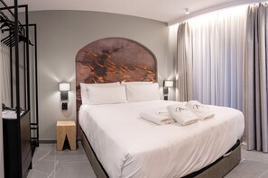Une chambre d’hôtel avec un lit, deux lampes de chevet, une table de chevet, un tabouret en bois et une grande fenêtre avec des rideaux.
