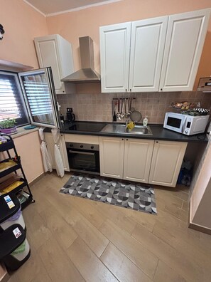 Cocina compacta con armarios blancos, microondas, horno y ventana con persianas.