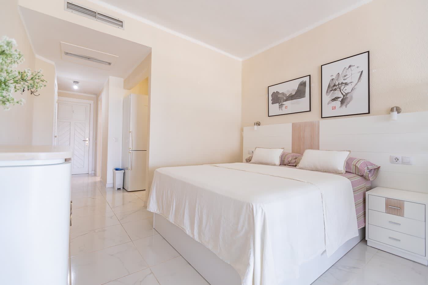 Quarto de hotel com cama, mesas de cabeceira, guarda-roupa e uma porta que dá para outro cômodo.