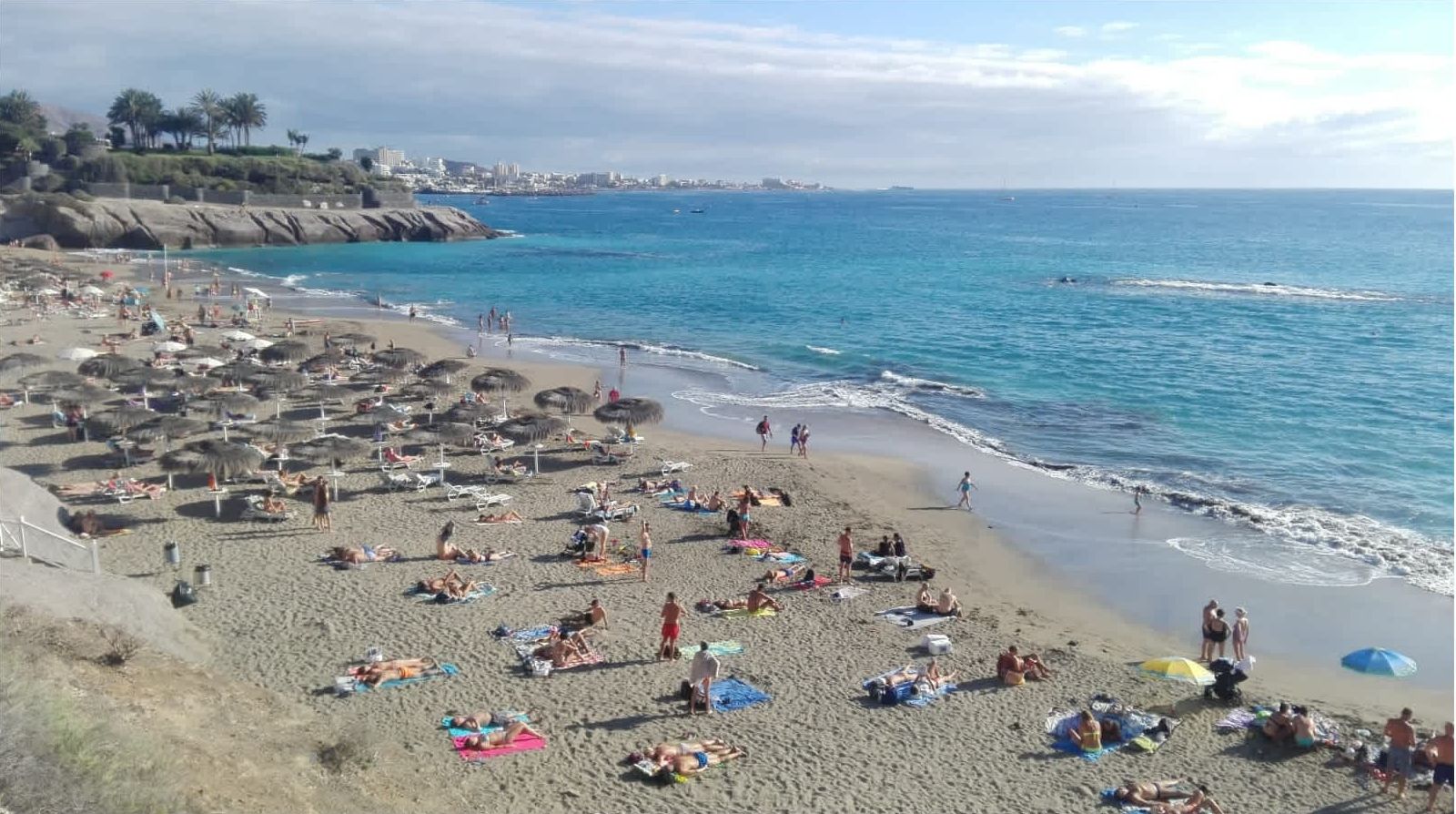 Uma praia com vários espreguiçadeiras e guarda-sóis, um litoral rochoso e uma vista ampla do oceano.