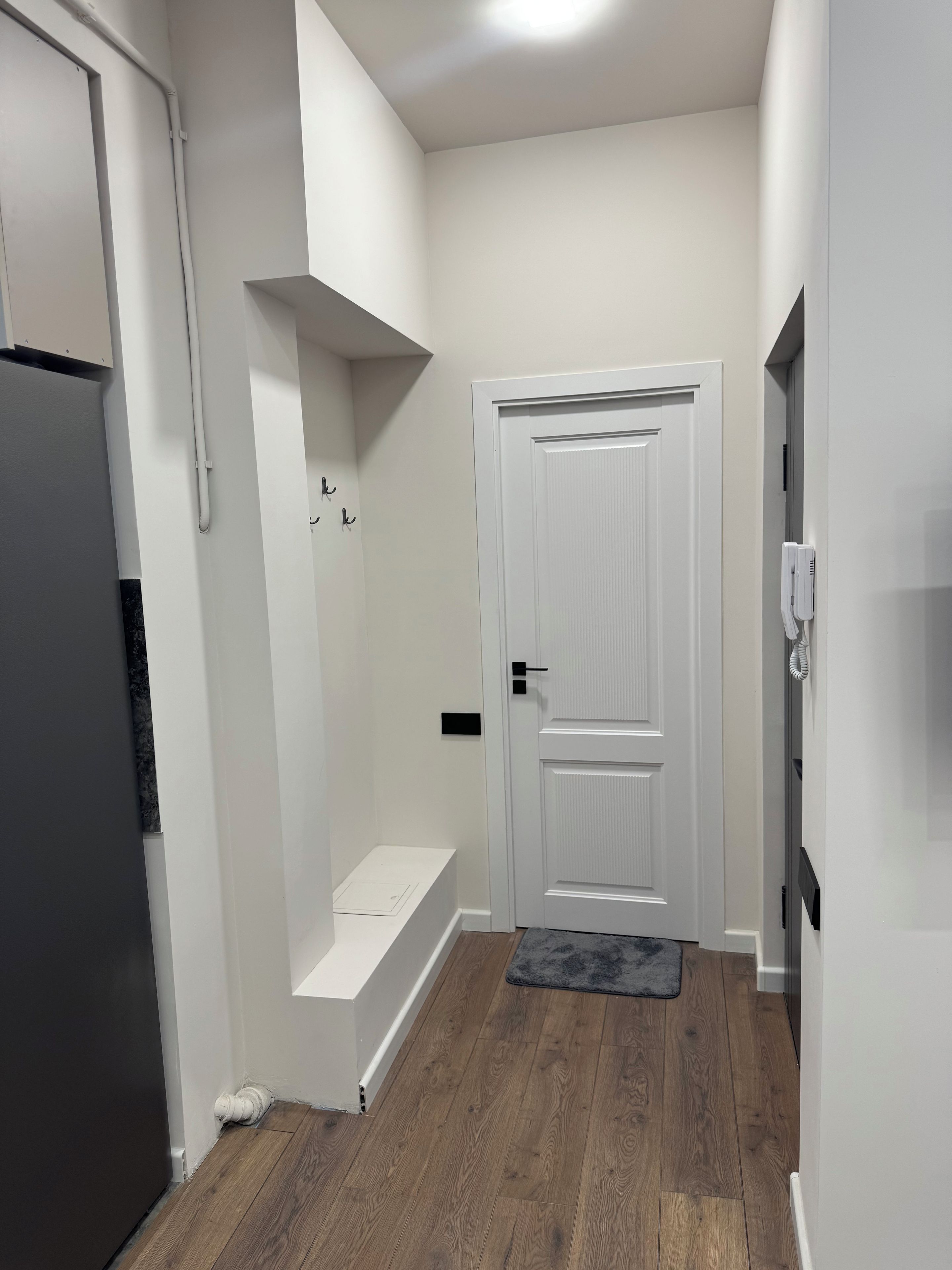 A narrow hallway with a white door, a white shelf, and a grey rug on a wooden floor.